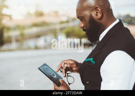 A mature gorgeous hairless black man entrepreneur with a beard and in an elegant costume is using outdoors a futuristic gadget, interacting with a glo Stock Photo