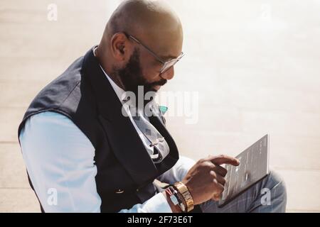 A mature bald bearded African businessman in an elegant outfit and glasses is using on the street a futuristic tablet pc with a glowing user interface Stock Photo