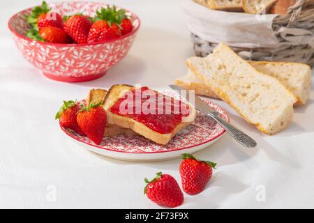 Juicy strawberry jam on slices of toast on a plate, with out of focus bowl of strawberries and basket of bread at the back. Selective focus. Stock Photo