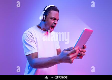 Online Gaming. Angry African Man In Wireless Headphones Using Digital Tablet Stock Photo
