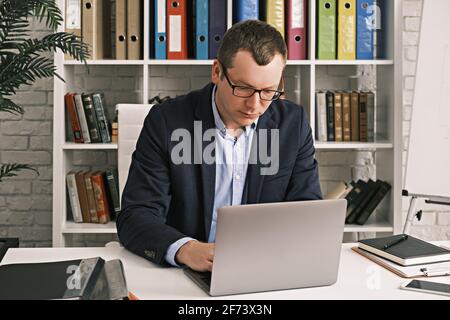Serious, focused man in glasses and business suit is typing something using laptop while sitting in light home office on the background of rack with Stock Photo