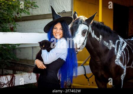 A girl dressed as a witch holds a black cat in her arms and stands by a corral on a farm next to a horse