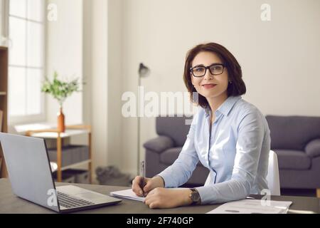 Portrait of a smiling middle aged businesswoman with glasses sitting at an office desk at work. Stock Photo