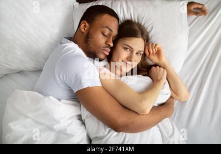 Young black guy sleeping and hugging his beautiful Caucasian girlfriend in bed, top view Stock Photo