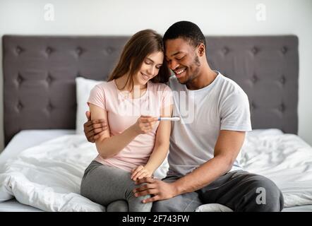 Portrait of happy multiracial couple holding positive pregnancy test, sitting on bed, hugging and enjoying good news Stock Photo