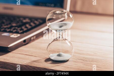 Hourglass on a light wooden table with a laptop or computer included. Time slips through my fingers. The concept of saving money and time. Stock Photo