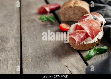 Italian cuisine concept. Crusty bread toast with cream cheese and thinly sliced ham, cherry tomatos, basil leaves branches, kitchen towel on wooden ba Stock Photo