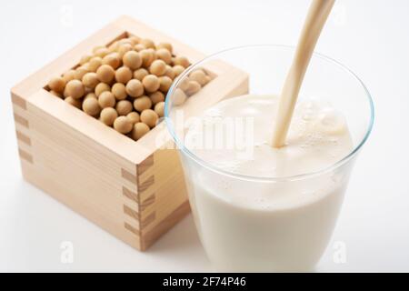 Pour soy milk into a glass placed on a white background. Soybeans in a Masu in the background Stock Photo