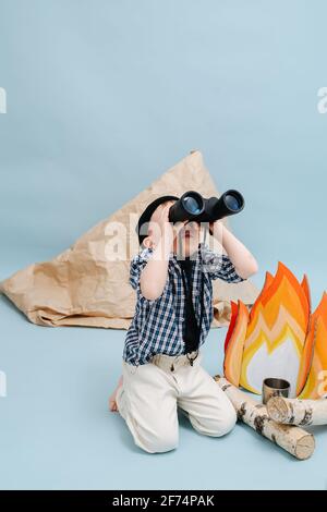 Pretending boy sitting on her knees next to a fake campfire and a paper tent, looking up through binoculars. Over blue background Stock Photo
