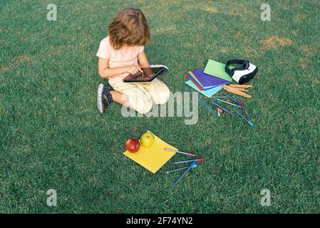 Child study exam outside. Funny kid little student boy with tablet, Sit on lawn in park, studying at School backyard. Stock Photo