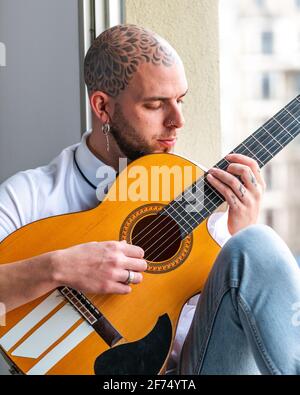 Pensive male with tattooed bald head in casual clothes sitting on windowsill and playing guitar in daytime Stock Photo
