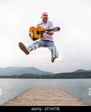 Full length of bearded guy in casual outfit jumping with guitar on wooden pier near lake with green mountains on background Stock Photo