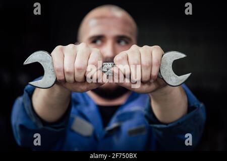 Faceless male mechanic in blue overall covering face with metal spanner in hands looking away on black background Stock Photo