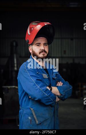 Serious bearded workman wearing protective hardhat and blue overall while standing in garage and looking at camera with crossed arms Stock Photo