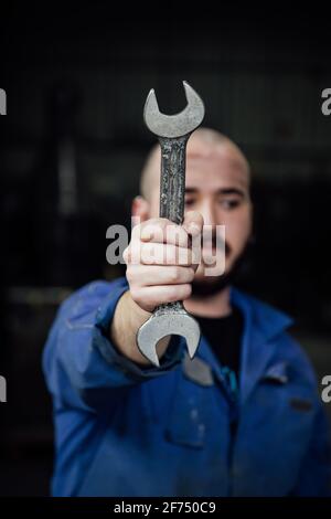 Male mechanic in blue overall covering face with metal spanner in hands looking away on black background Stock Photo