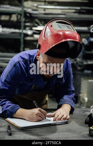 Serious bearded male worker in hardhat and overall taking notes in notebook with pen on table near metal constructions in light garage Stock Photo