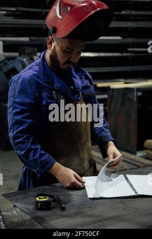 Serious bearded male worker in hardhat and looking notes in notebook with pen on table near metal constructions in light garage Stock Photo