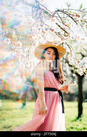 Peaceful ethnic female in straw hat and dress standing under blooming fragrant flowers on tree branches in orchard Stock Photo