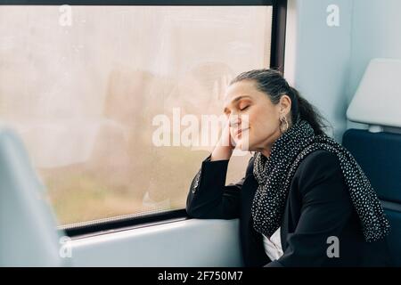 Side view of middle aged female with closed eyes leaning on hand while napping on passenger seat near window during ride in wagon Stock Photo