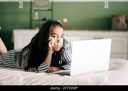 Side view of focused mature self employed Hispanic Woman with long dark hair in casual clothes lying on bed with laptop during online work at home Stock Photo