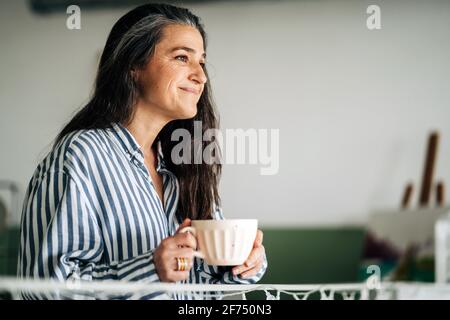 Side view of smiling middle aged female with cup of hot beverage looking into distance while sitting in room with paintings and easel on blurred backg Stock Photo