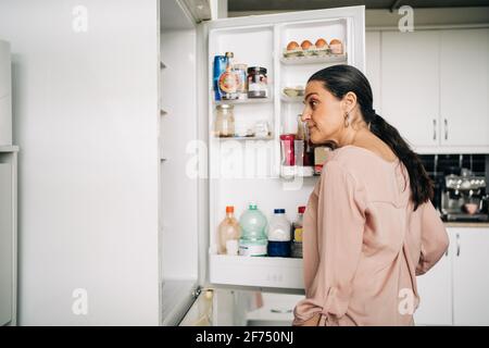 Side view of calm female with ponytail opening door of refrigerator with various products while standing in kitchen with white cupboards Stock Photo