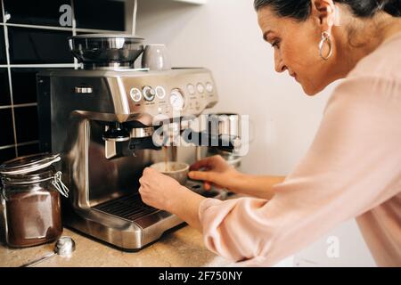 Side view of concentrated middle aged female preparing fresh coffee while using modern coffee maker at kitchen counter Stock Photo