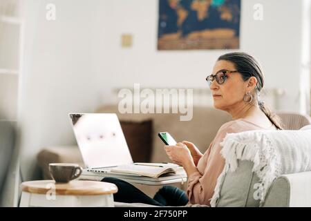 Side view of concentrated middle aged female browsing on smartphone while working on laptop in light room at home Stock Photo