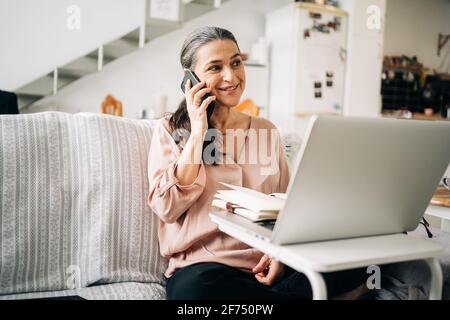 Concentrated middle aged female having phone call and taking notes in notepad while looking at screen of laptop in light room at home Stock Photo