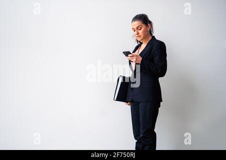 Serious middle aged female entrepreneur with ponytail wearing black suit text messaging on cellphone while standing on white background with folder Stock Photo