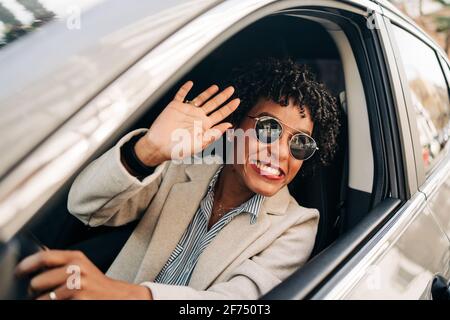 Cheerful African American female in stylish sunglasses and fashionable outfit smiling and waving hand in automobile Stock Photo