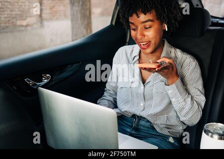 Busy young African American female recording voice message on mobile phone and working with netbook in automobile on backseat Stock Photo