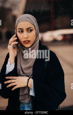Worried ethnic female in hijab standing in city street and having conversation on mobile phone while looking away Stock Photo