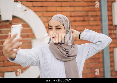 Young smiling Muslim female in traditional hijab standing on street and taking self shot on smartphone Stock Photo