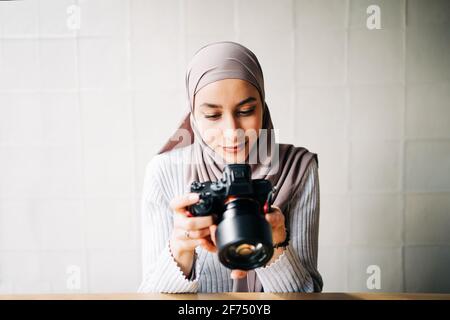 Focused Muslim female photographer sitting at table and looking through photos on professional camera while working remotely in cafe Stock Photo