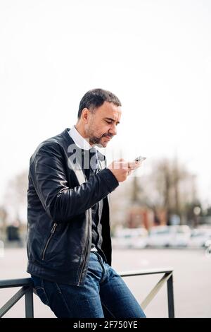Side view of middle aged ethnic male entrepreneur text messaging on cellphone while sitting on fence in city Stock Photo