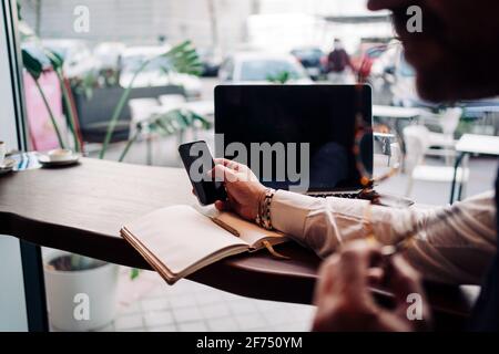 Side view of crop anonymous male executive touching black screen on cellphone at table with netbook in cafe Stock Photo