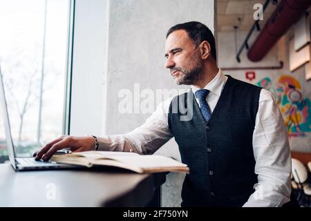 Mature Hispanic male entrepreneur in formal clothes working on netbook at cafe table with journal Stock Photo