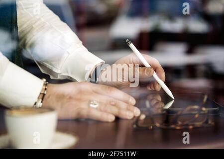 Through glass wall view of crop anonymous male executive using stylus and cellphone at cafeteria table Stock Photo