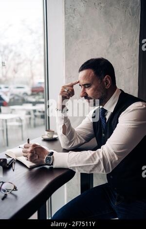 Side view of pensive mature Hispanic male entrepreneur touching forehead at table with agenda in cafeteria Stock Photo