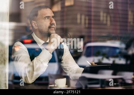 Thoughtful middle aged ethnic male entrepreneur touching mouth with glasses while using tablet behind glass wall in cafeteria Stock Photo