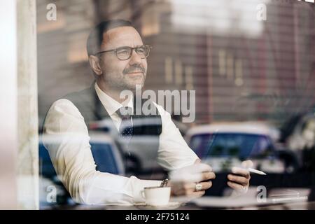 Thoughtful middle aged ethnic male entrepreneur using tablet behind glass wall in cafeteria Stock Photo
