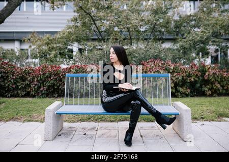 Stylish female entrepreneur sitting on bench and taking notes in organizer while working in urban park Stock Photo