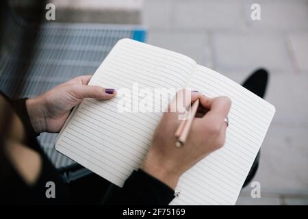High angle of crop unrecognizable female entrepreneur sitting on bench in city and writing business plan in notebook Stock Photo