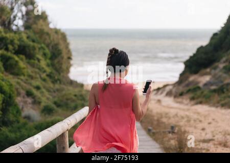Back view of anonymous female in pink silky blouse using mobile phone with black screen on coast of sea Stock Photo