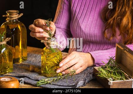 Crop anonymous lady in purple sweater showing essential oil glass bottles with herbs sprigs with green leaves near cloth on table Stock Photo