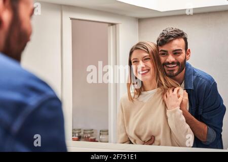 Crop cheerful young Hispanic man embracing smiling girlfriend while standing together in front of mirror Stock Photo