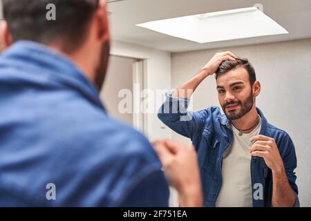 Side view of concentrated young bearded ethnic guy in stylish clothes touching hair while looking in mirror in modern bathroom Stock Photo