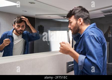 Side view of concentrated young bearded ethnic guy in stylish clothes touching hair while looking in mirror in modern bathroom Stock Photo