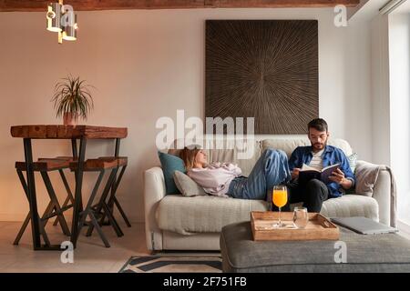 Tired young female in casual clothes sleeping on comfortable sofa near focused ethnic boyfriend reading book in daylight Stock Photo